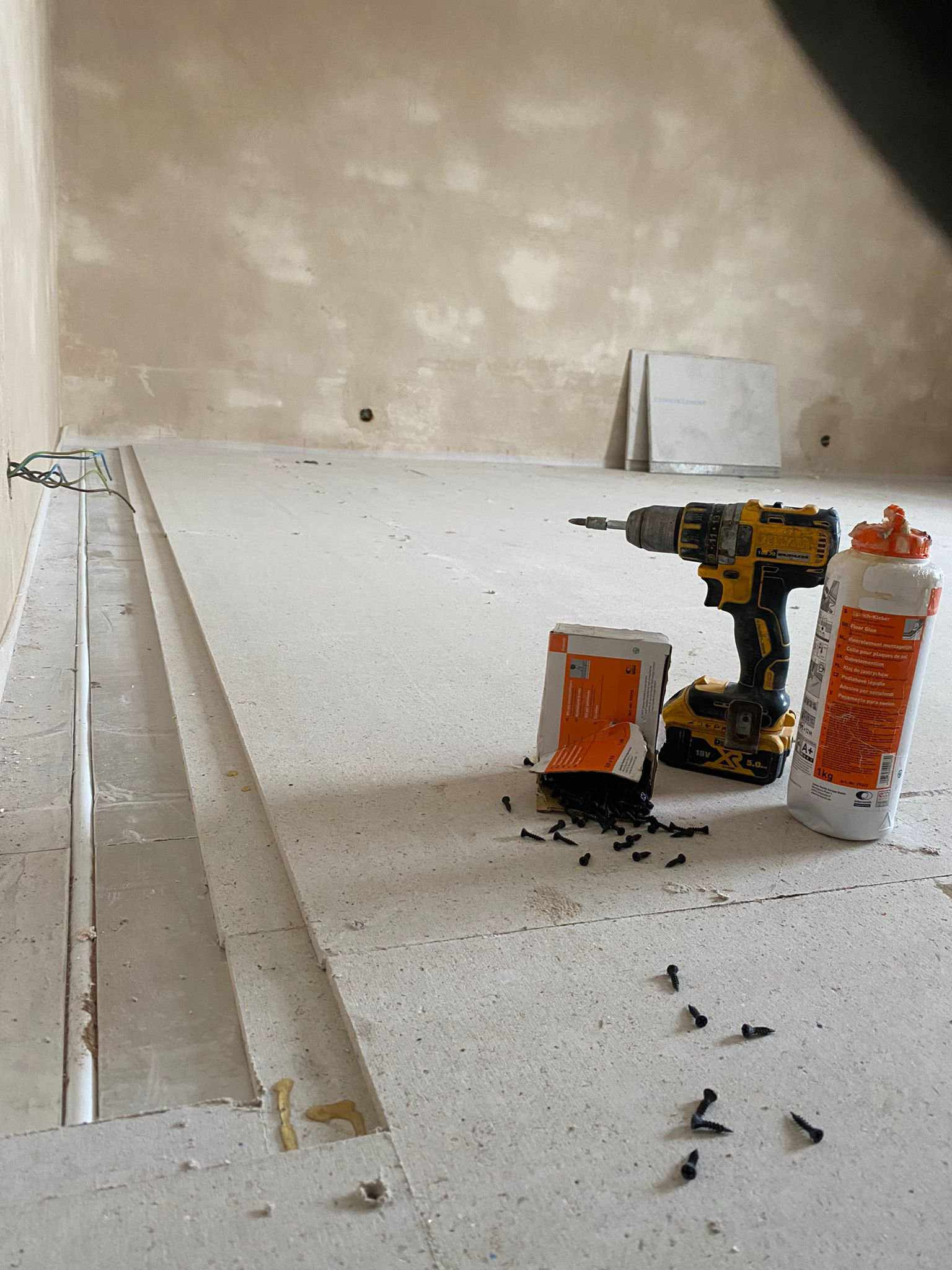 Przygotowanie do montażu ogrzewania podłogowego: płyty izolacyjne podłogowe, wkręty, klej i wiertarko-wkrętarka DeWalt na surowej posadzce w trakcie prac instalacyjnych.