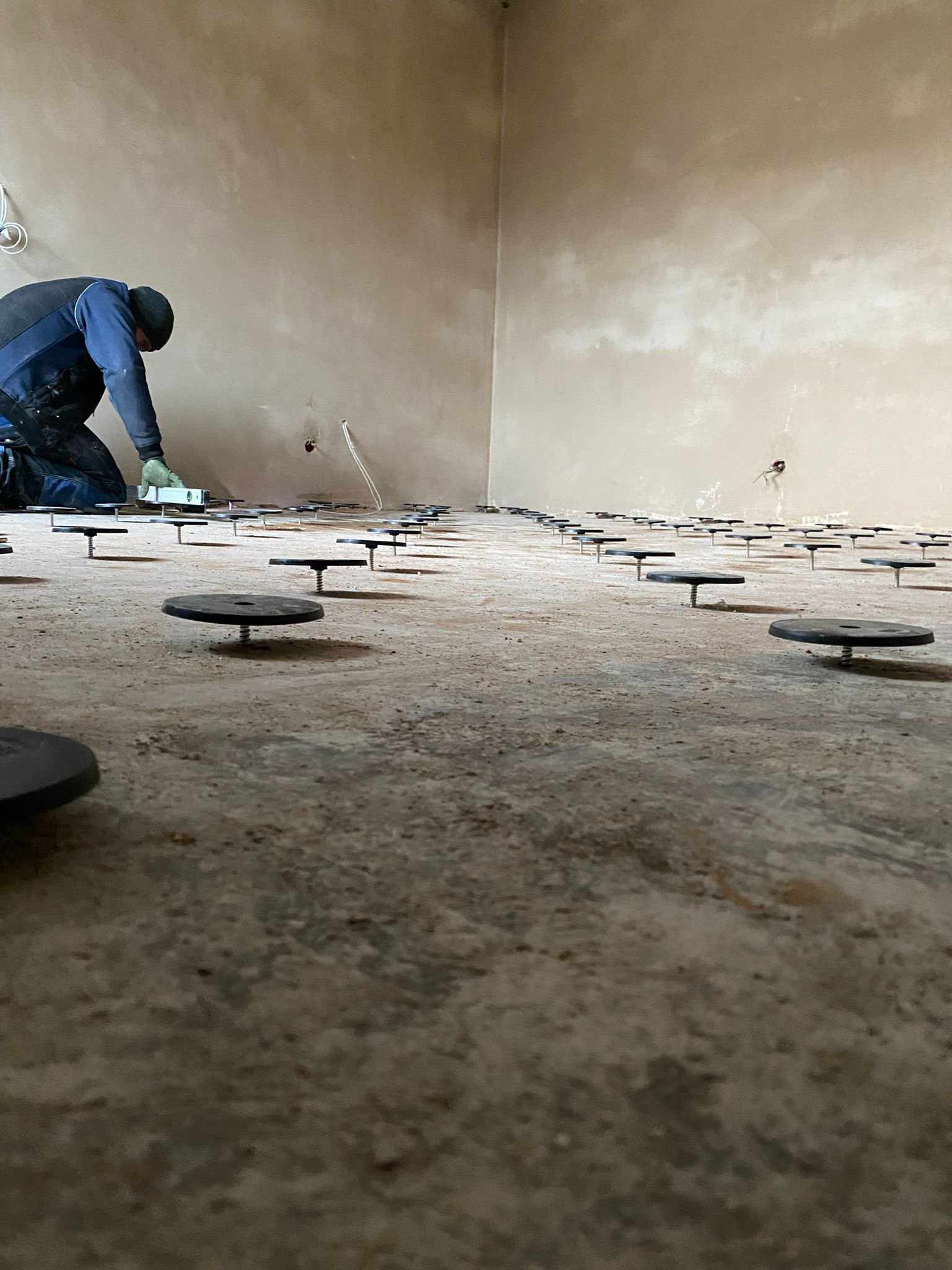 Instalacja ogrzewania podłogowego: widok z niskiego kąta na przygotowany beton z rozmieszczonymi wspornikami poziomującymi, instalator z poziomicą w tle.