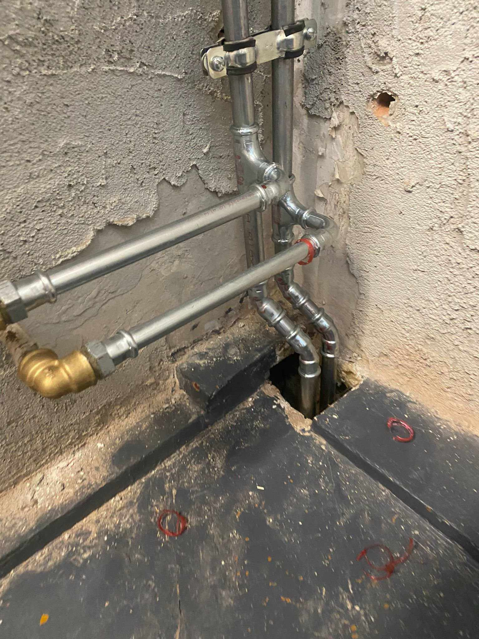 Fragment instalacji wodnej z rur stalowych i mosiężnych, widoczne połączenia gwintowane i obejmy mocujące, surowy stan wykończenia ściany i podłogi.