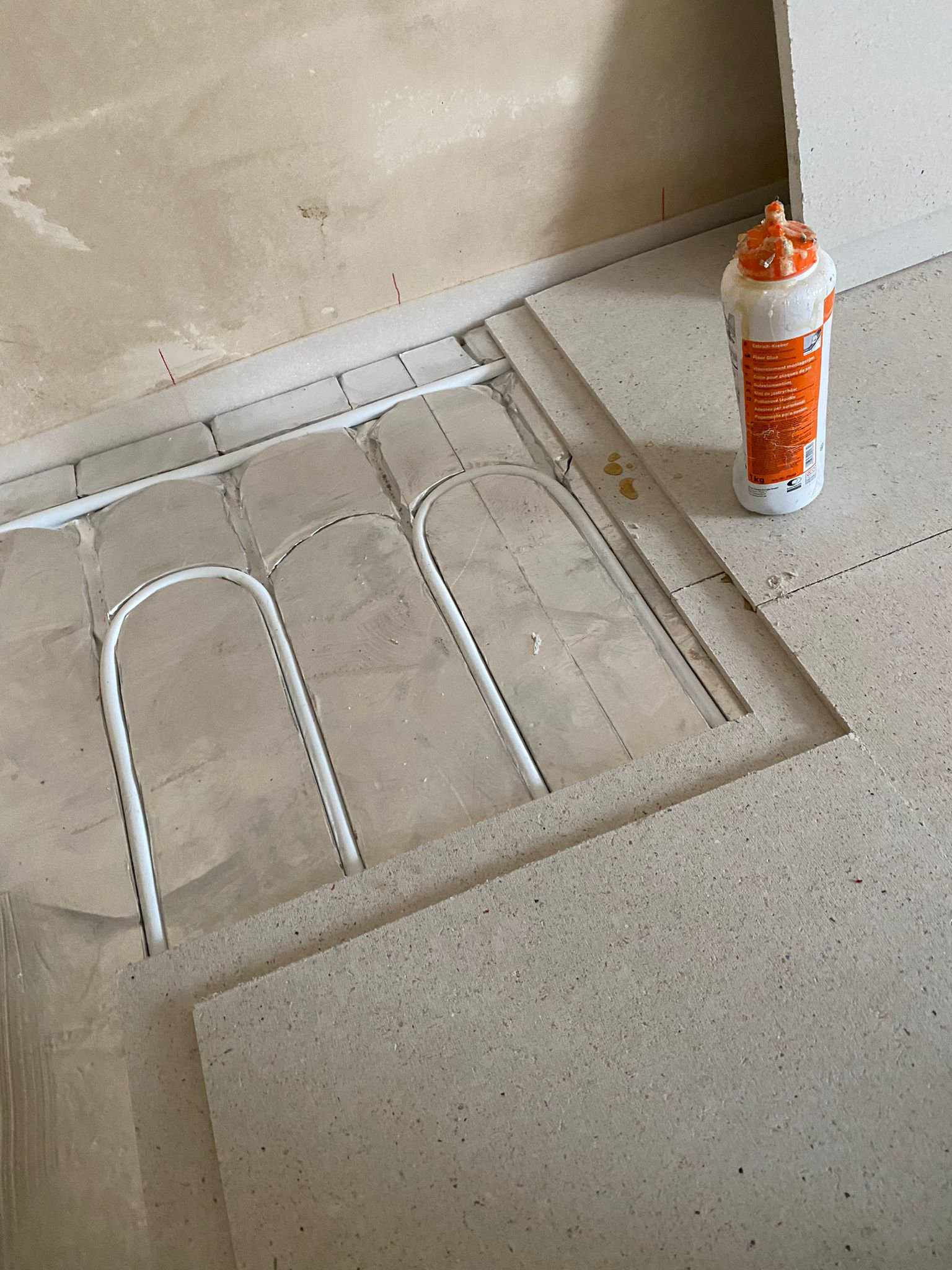 Białe rurki systemu grzewczego ułożone na srebrnej folii izolacyjnej, widoczne wycięte płyty podłogowe i butelka kleju montażowego w tle.