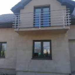 Szara elewacja domu z balkonem zabezpieczonym balustradą nierdzewną z poziomych profili, widoczne okna z szarą ramą oraz drzwi wejściowe z przeszkleniem.