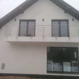 Mikołaj - Biały dom jednorodzinny z balkonem zabezpieczonym prostą, metalową konstrukcją, widoczne okna z ciemnymi ramami i fragment ogrodu w tle.