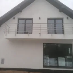 Biały dom jednorodzinny z balkonem zabezpieczonym prostą, metalową konstrukcją, widoczne okna z ciemnymi ramami i fragment ogrodu w tle.