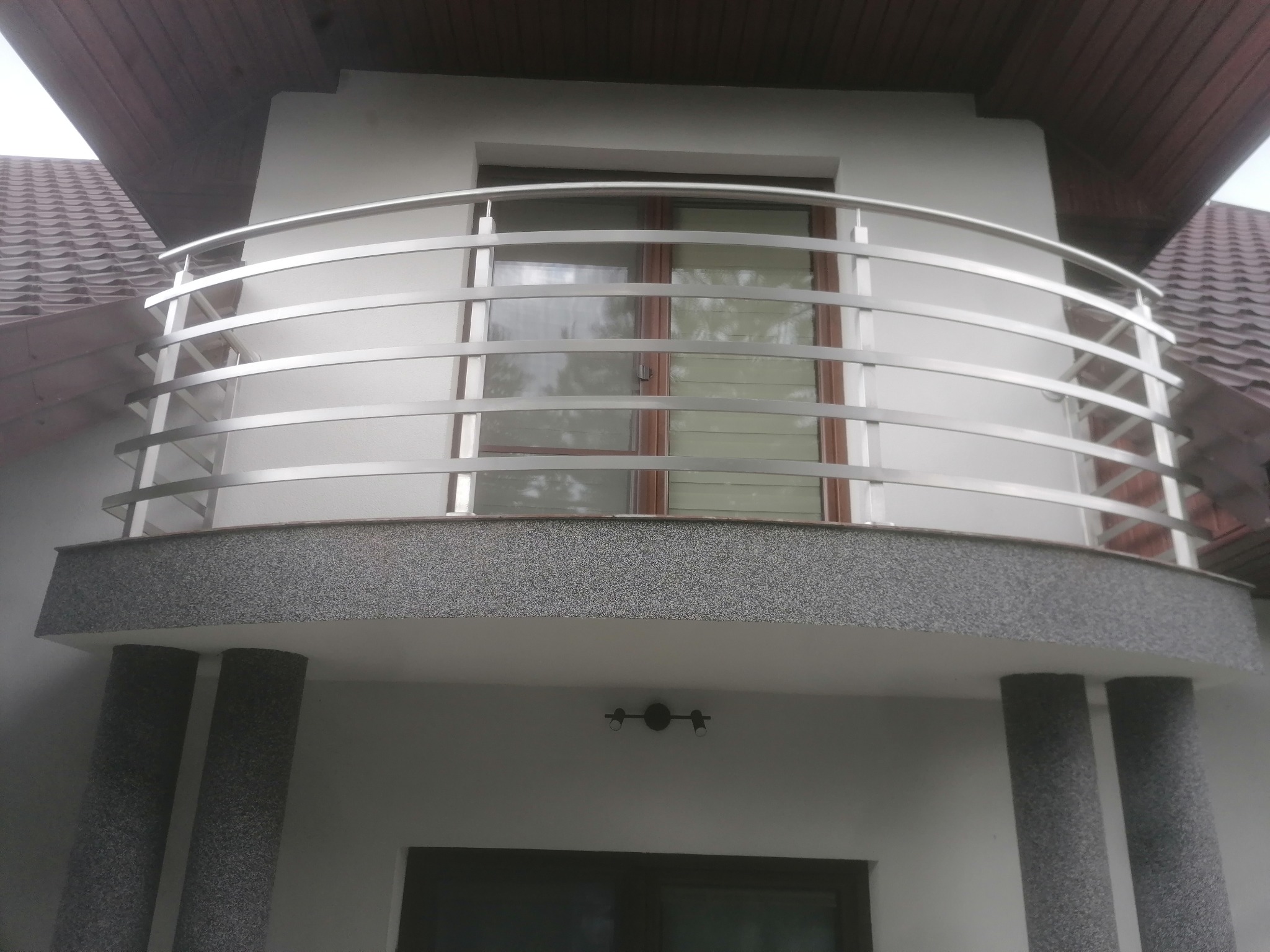 Balkon z zaokrągloną, metalową konstrukcją zabezpieczającą, widoczny z dołu, z elementami elewacji w tle.