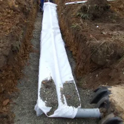 Wykop z rurą drenażową owiniętą białą geowłókniną, częściowo zasypaną żwirem, widoczne ściany wykopu z ziemi.