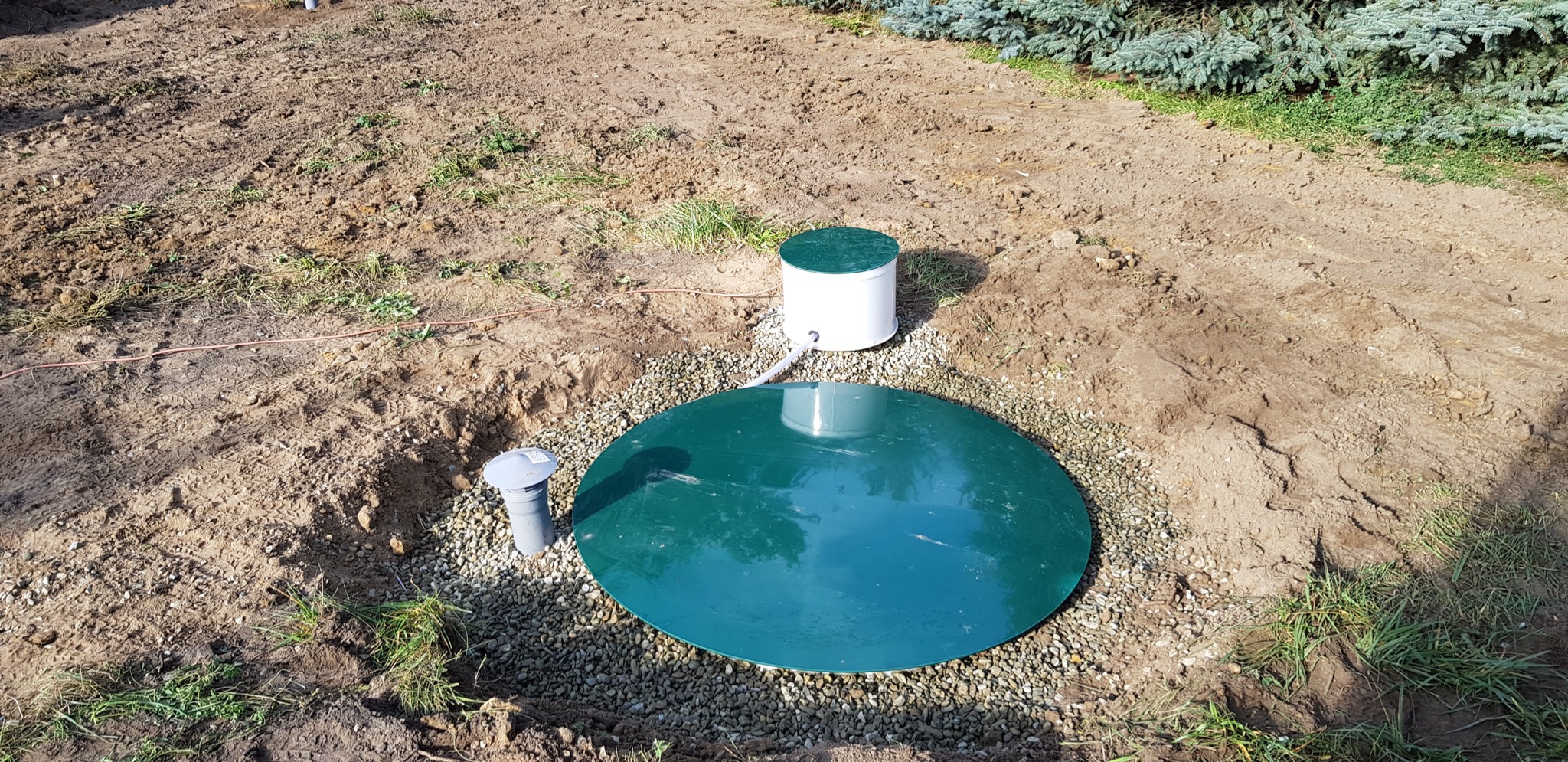 Zielona pokrywa zbiornika z szarego kamienia, biały cylinder z zieloną pokrywą i szary kominek wentylacyjny wystają z ziemi na tle ziemi i zieleni.