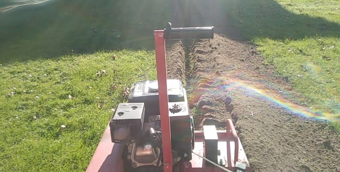 Koparka łańcuchowa tworzy wąski wykop w trawniku, widoczna tęcza spowodowana odbiciem światła na mokrej ziemi.