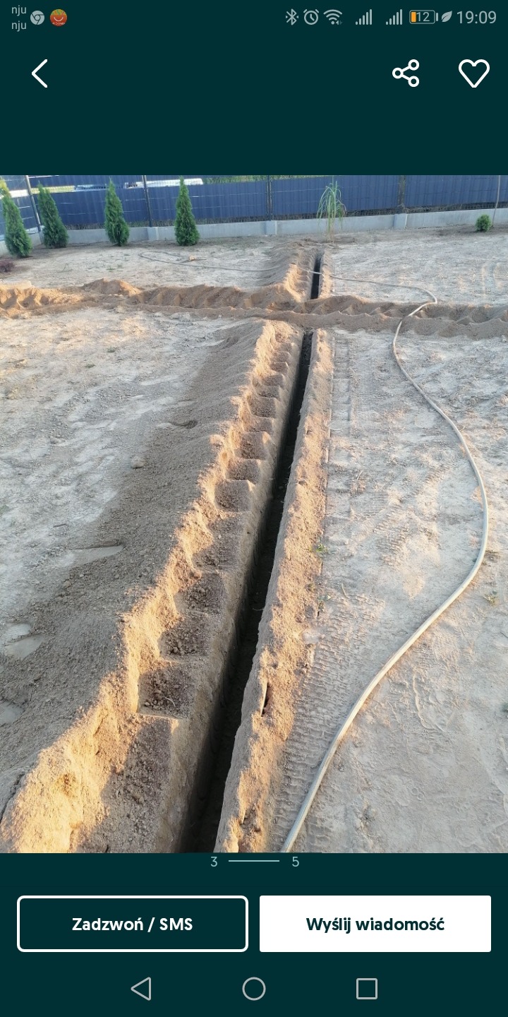 Wykopane wąskie rowy w piaszczystej ziemi, prawdopodobnie pod instalacje, z widocznym w tle ogrodzeniem i tujami.