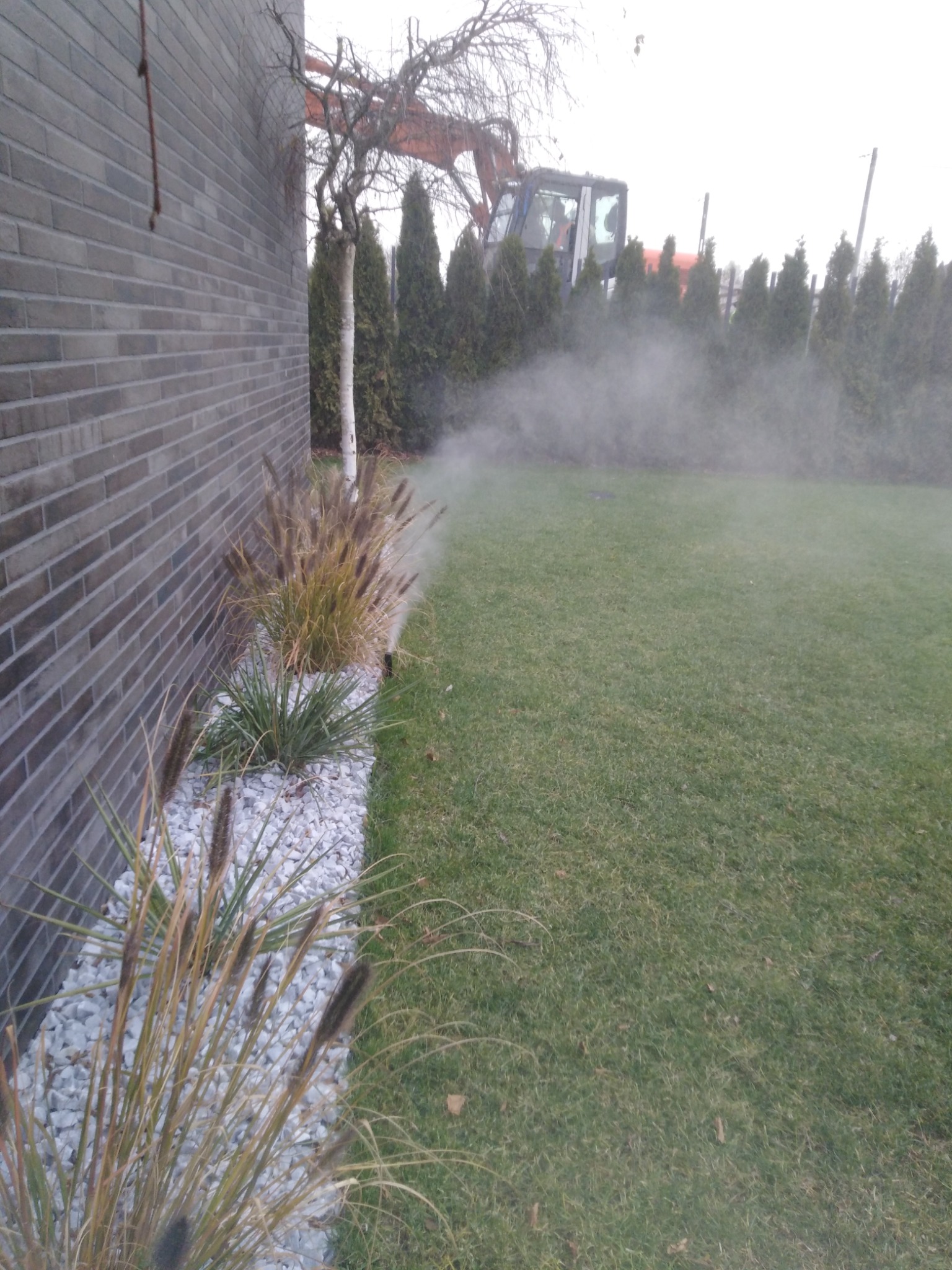 Automatyczne nawadnianie trawnika w ogrodzie, zraszacz rozpyla wodę, w tle widoczny budynek z cegły i koparka.