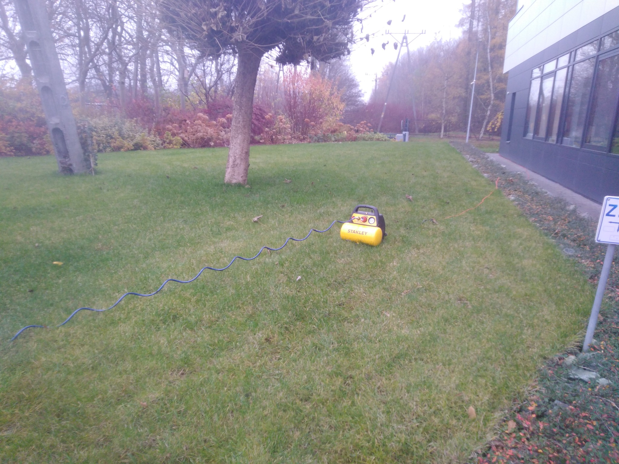 Żółty kompresor Stanley leży na trawniku obok budynku z ciemnymi oknami, widoczny kabel zasilający, drzewa w tle.