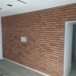 Ściana wykończona czerwoną cegłą klinkierową, widoczne przewody elektryczne wystające ze ściany i puszka instalacyjna, podłoga w trakcie prac wykończeniowych.