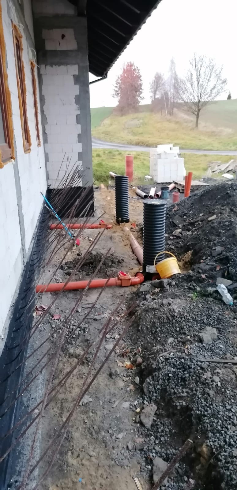 Instalacja rur kanalizacyjnych zewnętrznych w wykopie przy fundamentach budynku w trakcie budowy, widoczne czarne rury karbowane, pomarańczowe rury kanalizacyjne z kształtkami i żółte wiadro.