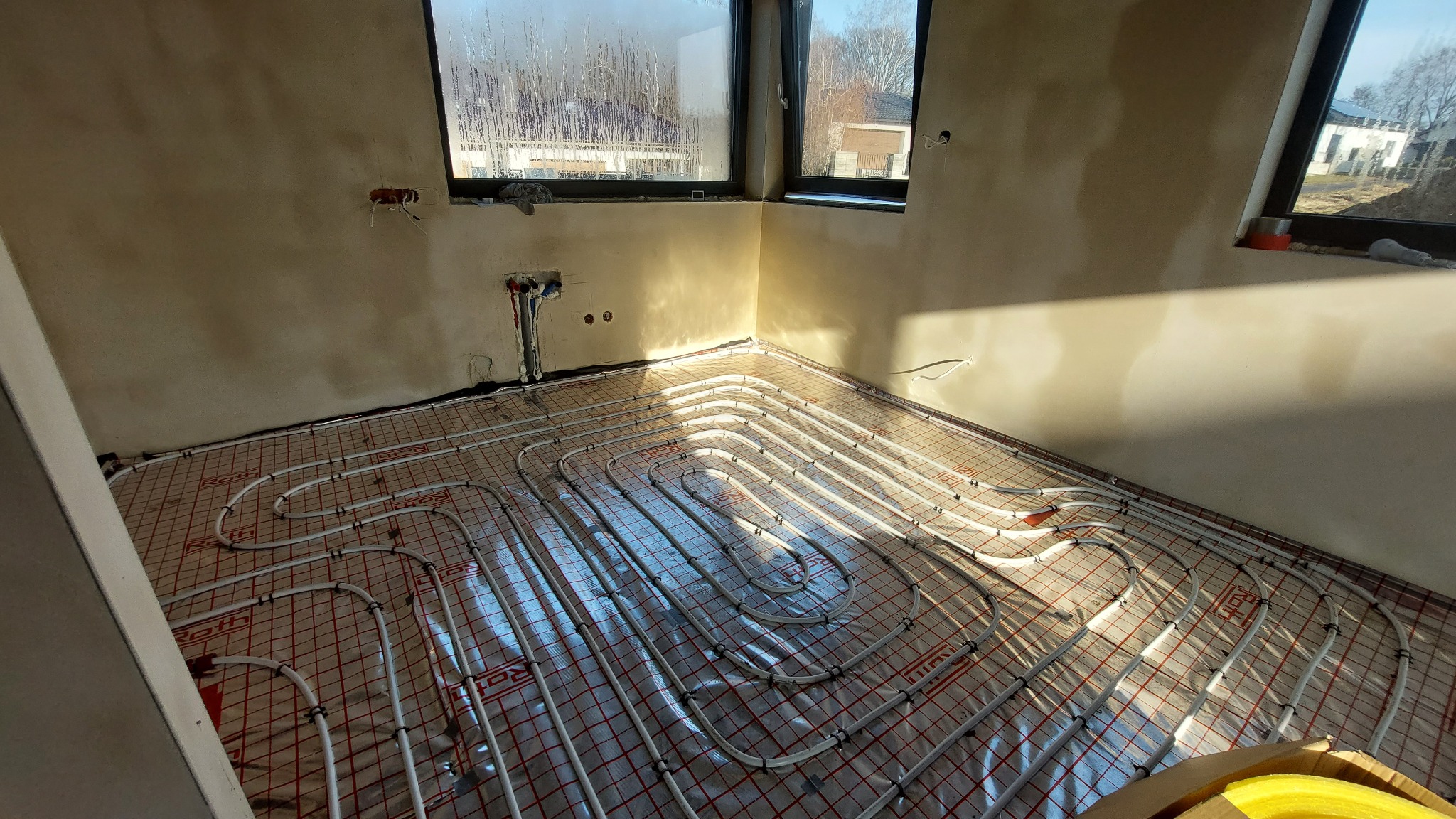 Wnętrze w trakcie montażu ogrzewania podłogowego: białe rurki układane na folii z czerwoną siatką, widoczne okna z refleksami światła słonecznego, surowe ściany w odcieniach beżu.