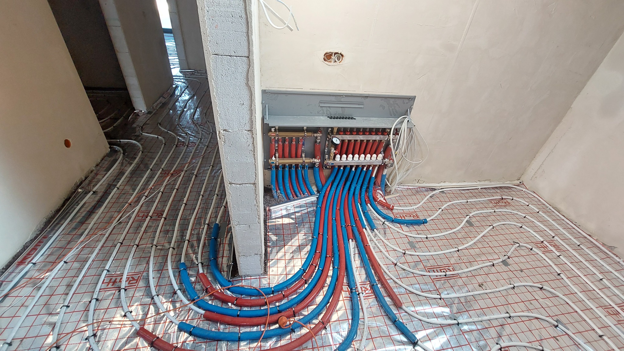 Rozłożona instalacja ogrzewania podłogowego z czerwono-niebieskimi rurami podłączonymi do rozdzielacza na ścianie, widok z góry w surowym stanie wykończenia.