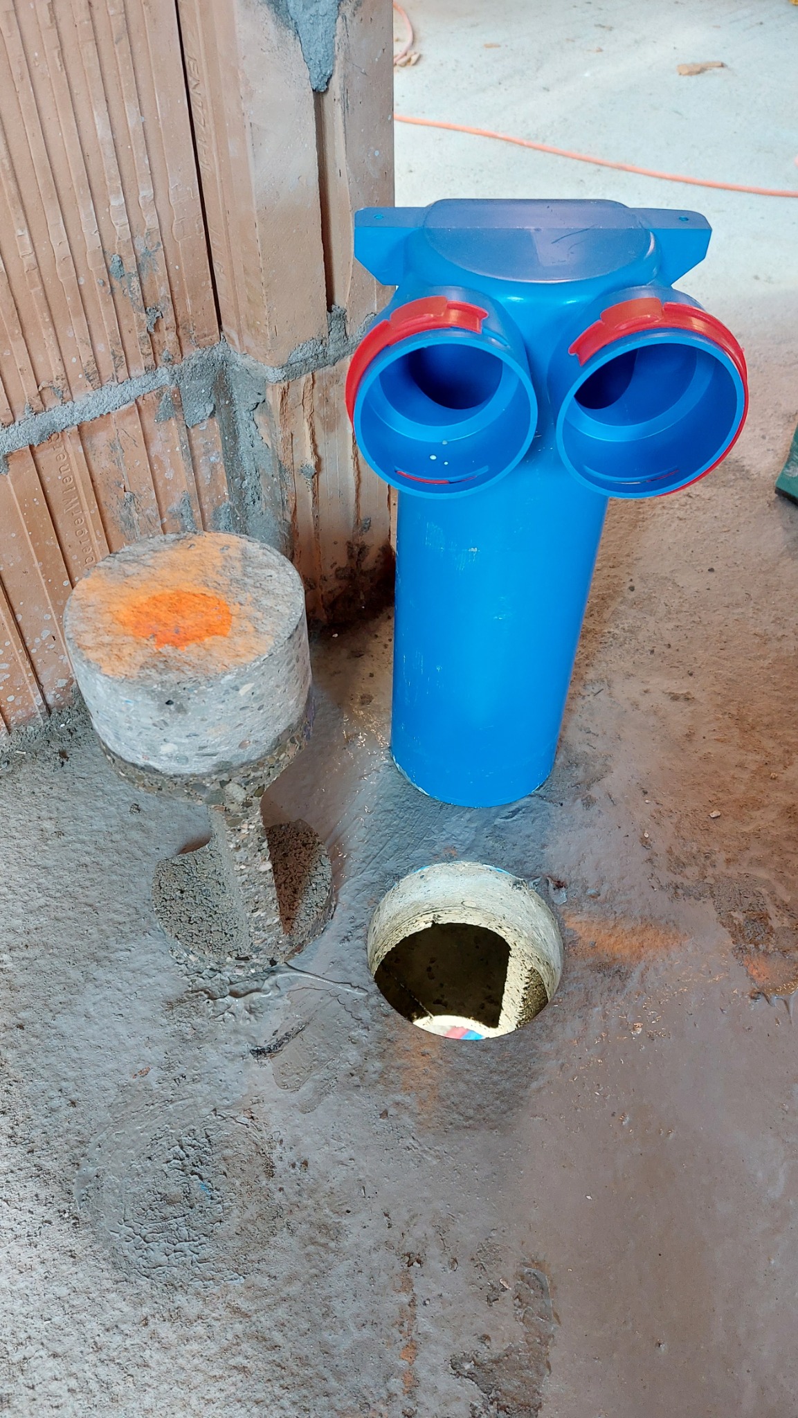 Niebieski rozdzielacz z rurami z czerwonymi elementami mocującymi, obok betonowy słupek z pomarańczowym oznaczeniem i wywiercony otwór w szarej, betonowej posadzce w surowym stanie.