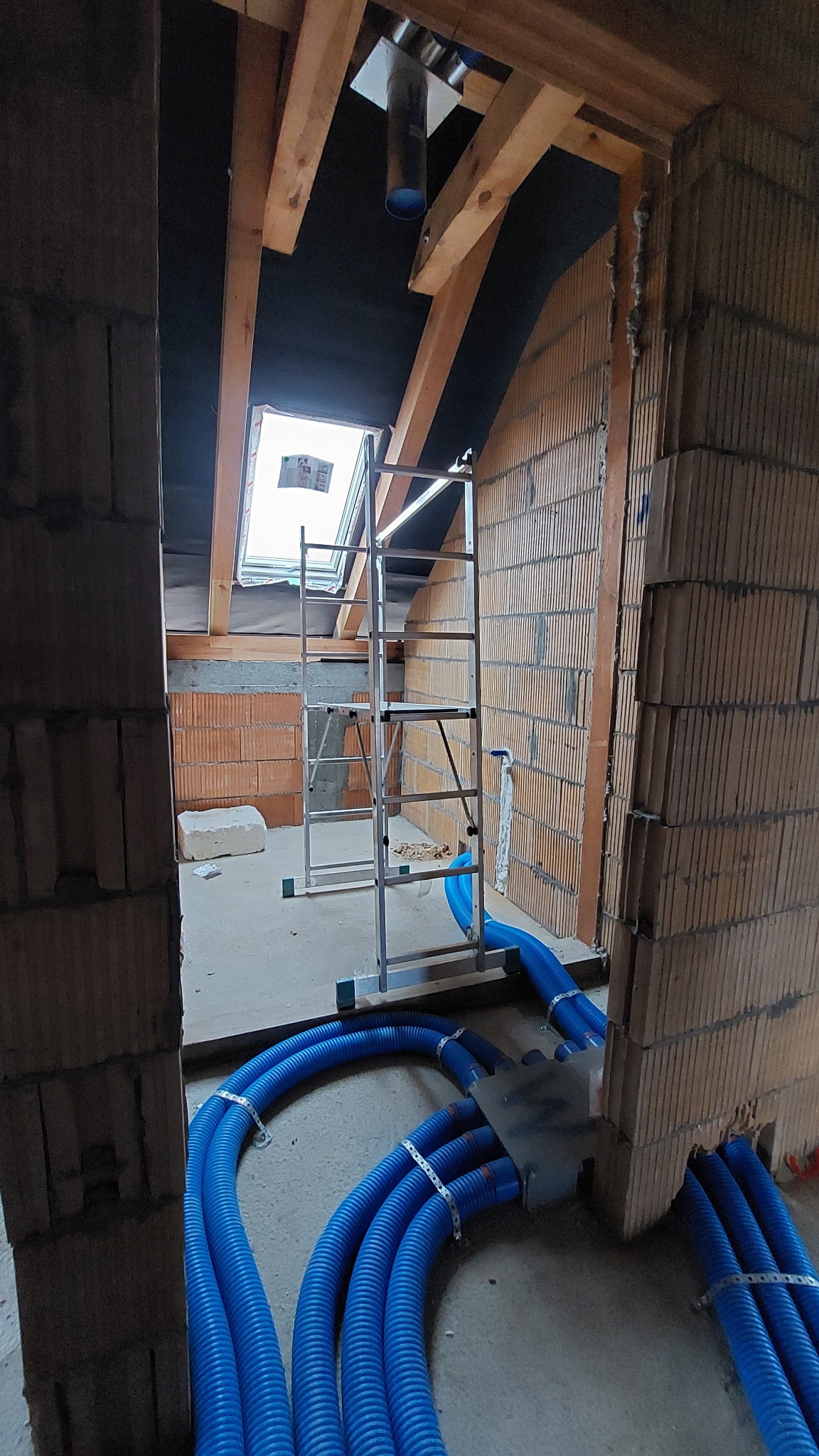 Niedokończone poddasze z widocznymi niebieskimi rurami instalacyjnymi, drabina aluminiowa ustawiona pod oknem dachowym, surowy stan wykończenia ścian z cegły.