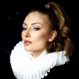 Portret kobiety z włosami upiętymi w wysoki kucyk, z delikatnym makijażem i w białym, plisowanym kołnierzu na czarnym tle.