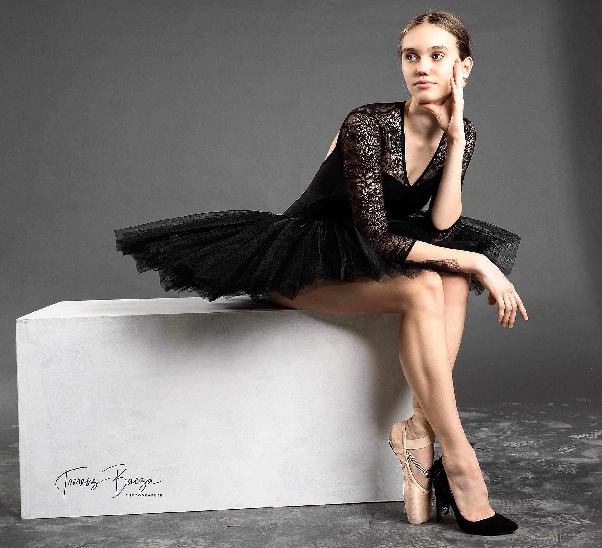 Młoda balerina w czarnej sukience z koronkowymi rękawami i pointach oraz czarnym szpilce pozuje siedząc na białym postumencie na szarym tle.