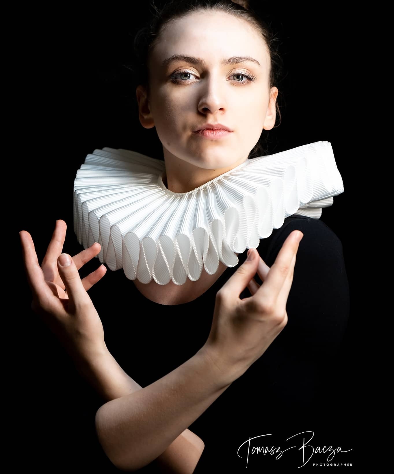 Portret kobiety w czarnej bluzce z białym, plisowanym kołnierzem, ręce skrzyżowane, ciemne tło, ostre światło.