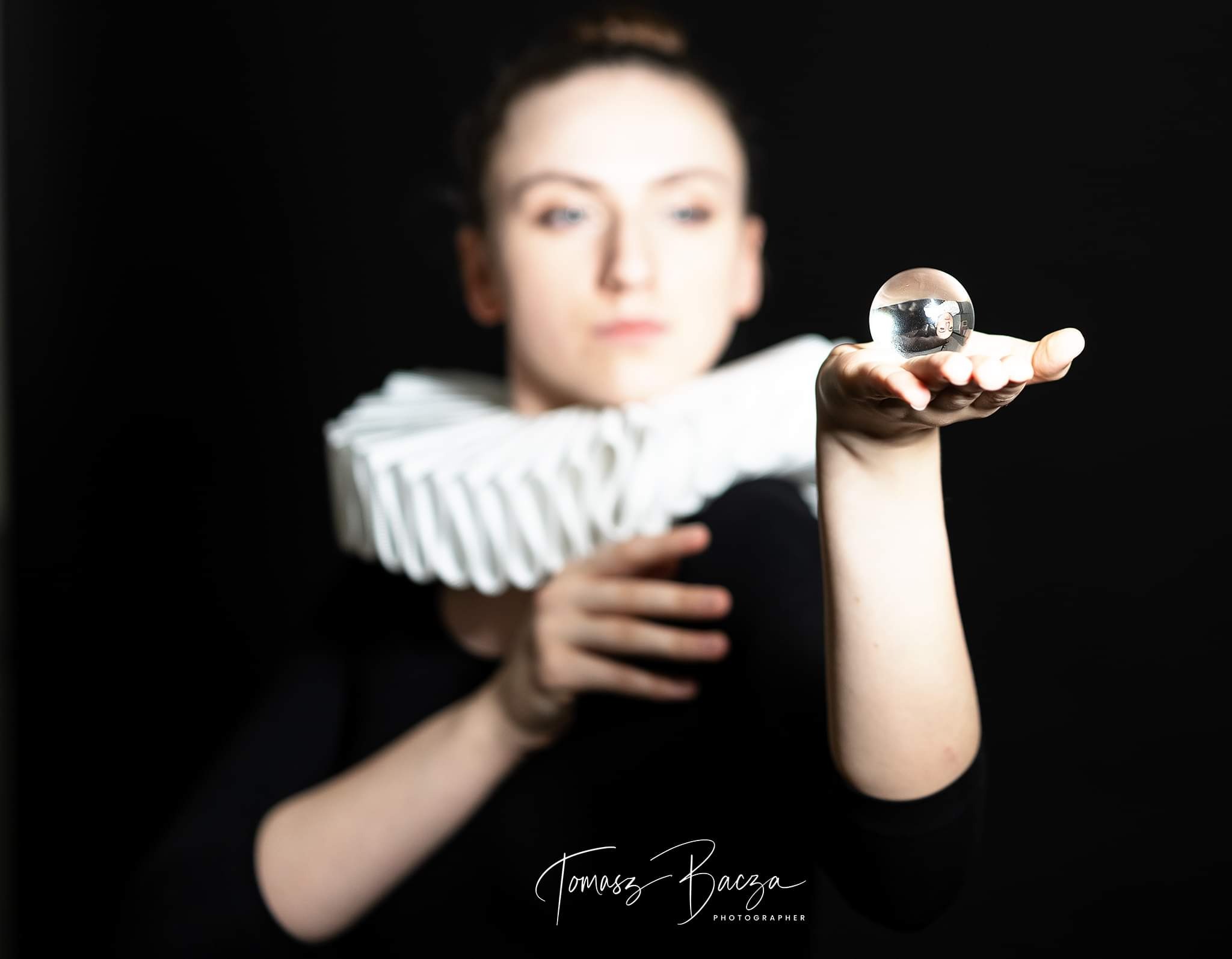 Kobieta z białym, plisowanym kołnierzem trzymająca szklaną kulę, w której odbija się jej twarz, na czarnym tle.
