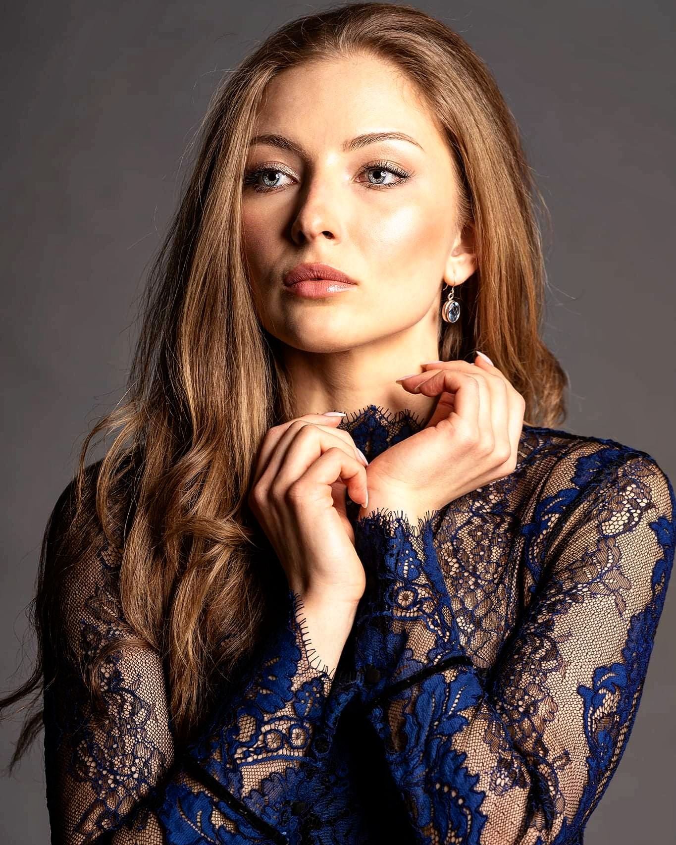 Portret kobiety w koronkowej sukience z niebieskimi wzorami, delikatny makijaż, subtelna biżuteria, dłonie splecione pod szyją, patrząca w górę.
