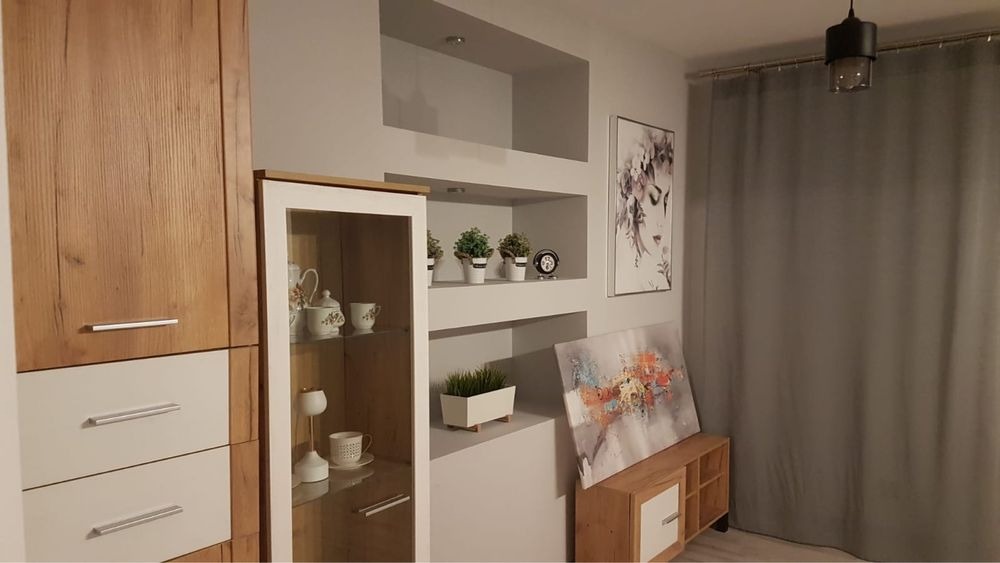 Nowoczesny salon z drewnianą zabudową meblową, szklaną witryną, wnękami w ścianie z dekoracjami i obrazami, oświetlony wiszącą lampą.