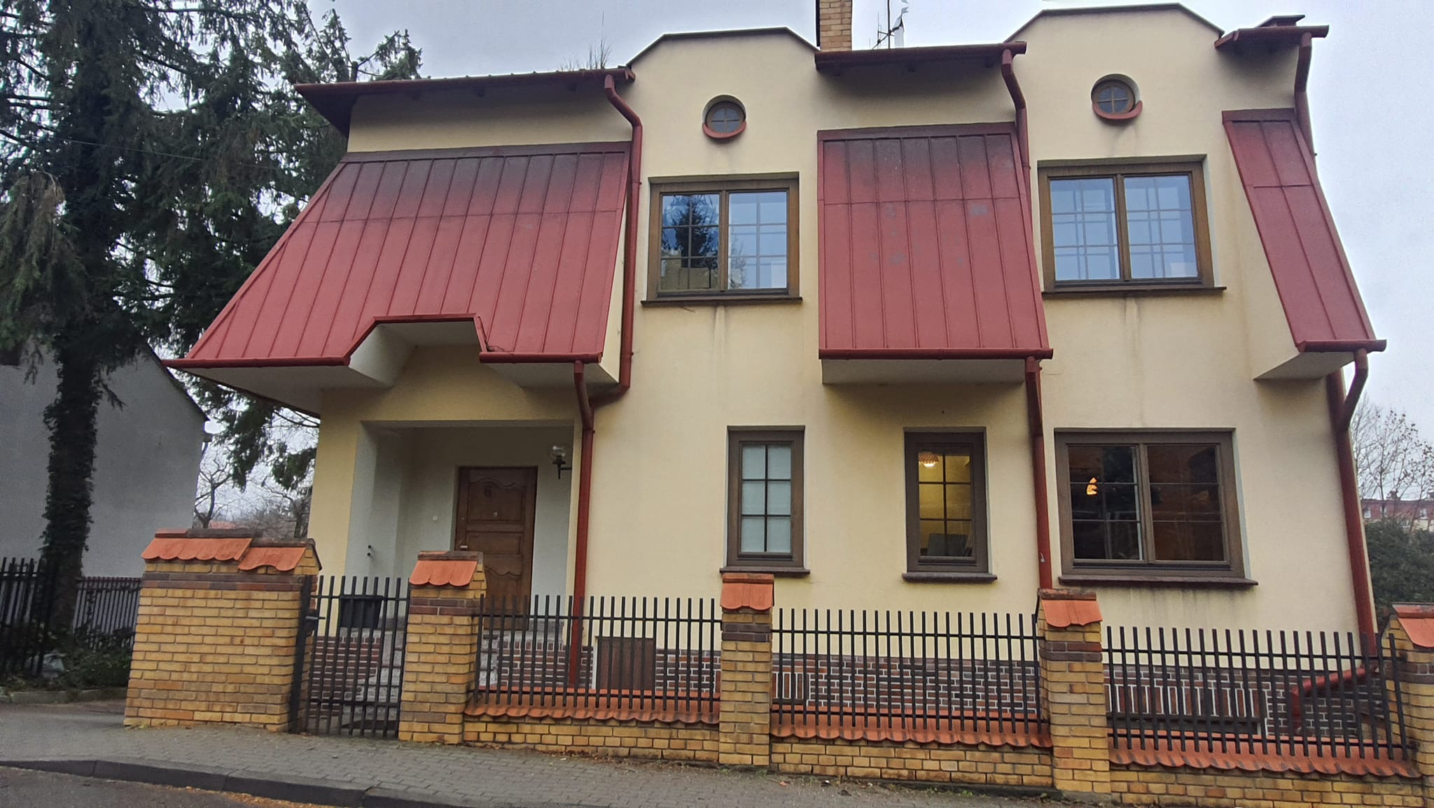 Dwukondygnacyjny dom z beżową elewacją, czerwonym dachem i ogrodzeniem z cegły i metalu, widziany z perspektywy ulicy w pochmurny dzień.