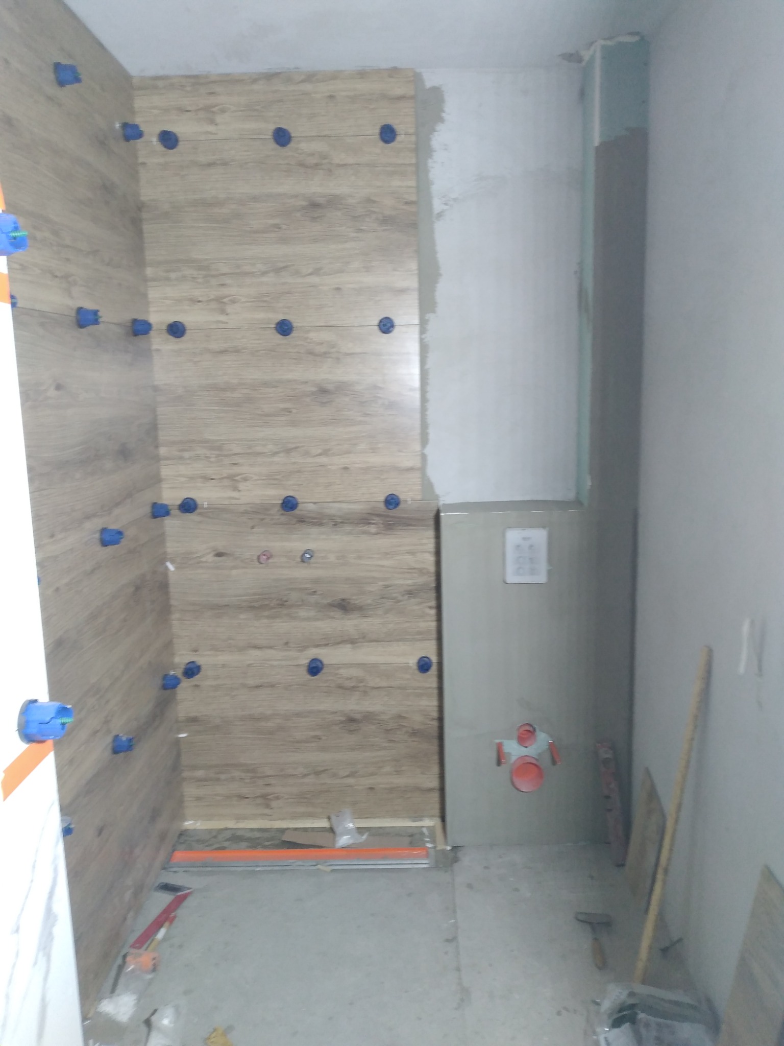 W trakcie układania płytek imitujących drewno na ścianie łazienki, widoczne niebieskie krzyżyki dystansowe i poziomica, surowy stan ścian w tle.