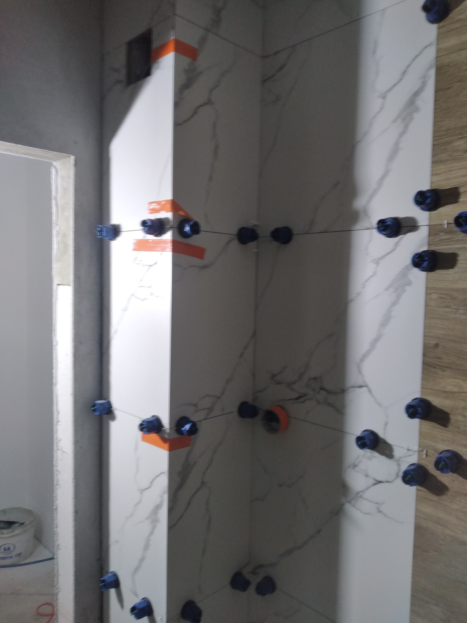 Układanie płytek imitujących marmur w rogu pomieszczenia, z użyciem pomarańczowej taśmy i niebieskich klipsów poziomujących.