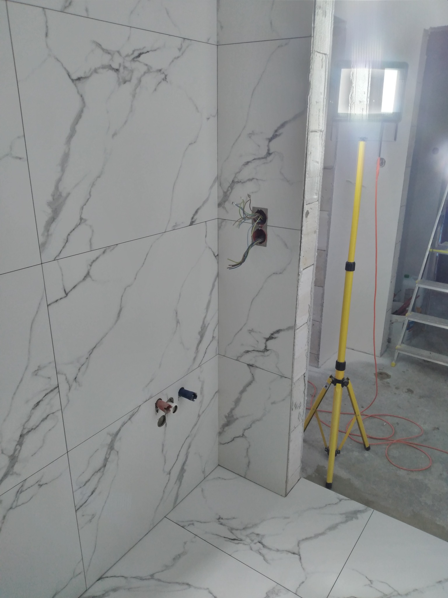 Wnętrze łazienki w trakcie prac wykończeniowych, widoczne białe płytki imitujące marmur na ścianach i podłodze, wystające przewody elektryczne, rury instalacyjne oraz żółty reflektor budowlany...
