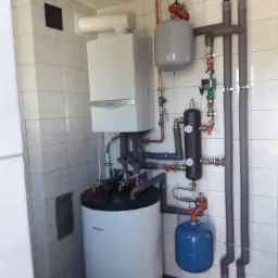 Zestaw instalacji grzewczej z kotłem gazowym i zasobnikiem ciepłej wody, widoczne rury miedziane i szare izolowane, manometry i zawory, całość zamontowana w pomieszczeniu wyłożonym białymi płytkami.