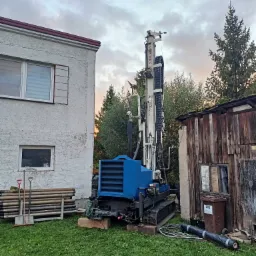 Wiertnica geologiczna na gąsienicach, ustawiona na trawniku obok budynku z widocznymi rurami wiertniczymi i narzędziami, w tle drewniana szopa i drzewa.