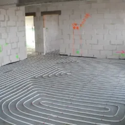 Rozłożone rury ogrzewania podłogowego na izolacji w surowym stanie budynku, widoczne oznaczenia na ścianach z betonu komórkowego.