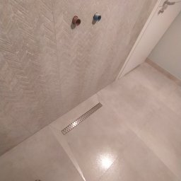 RoseGroup - Wykończona łazienka z beżowymi płytkami ułożonymi w jodełkę na ścianie i podłodze, widoczny odpływ liniowy oraz dwa wystające przyłącza wodne.