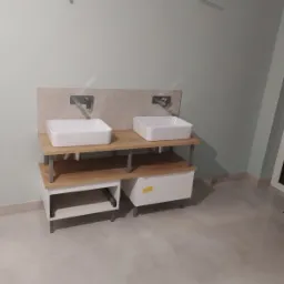 Dwie białe, prostokątne umywalki nablatowe z chromowanymi bateriami, zamontowane na drewnianym blacie wspartym na białych szafkach z szufladami, na tle jasnej ściany w łazience.