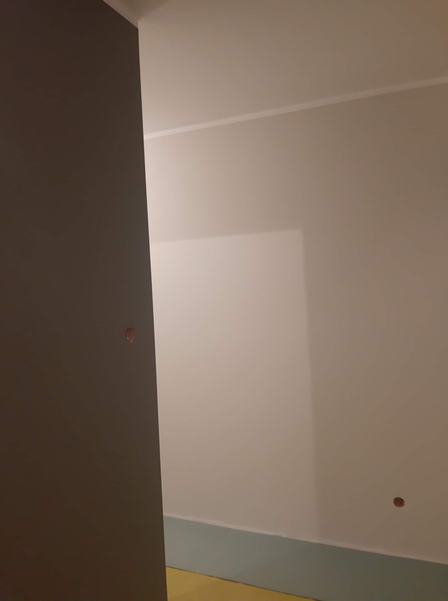 Fragment pokoju w trakcie remontu, widoczna ściana pomalowana na jasny kolor z widocznym otworem po kołku, dolna część ściany zabezpieczona niebieską folią malarską, obok ciemny fragment ściany.