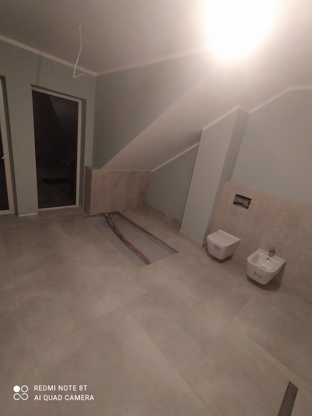 Łazienka w trakcie remontu w poddaszu, widoczne przygotowane miejsce na instalację prysznica, zamontowana toaleta i bidet, ściany w jasnych odcieniach szarości i beżu.