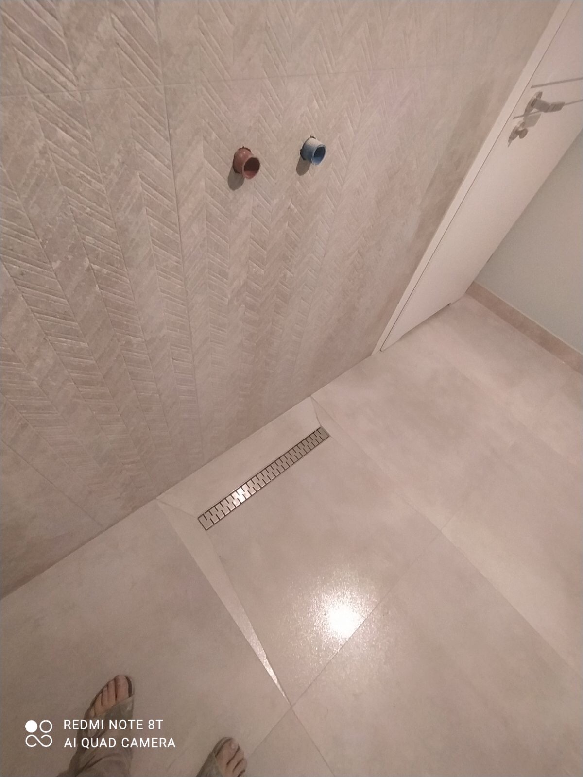 Wykończona łazienka z beżowymi płytkami ułożonymi w jodełkę na ścianie i podłodze, widoczny odpływ liniowy oraz dwa wystające przyłącza wodne.