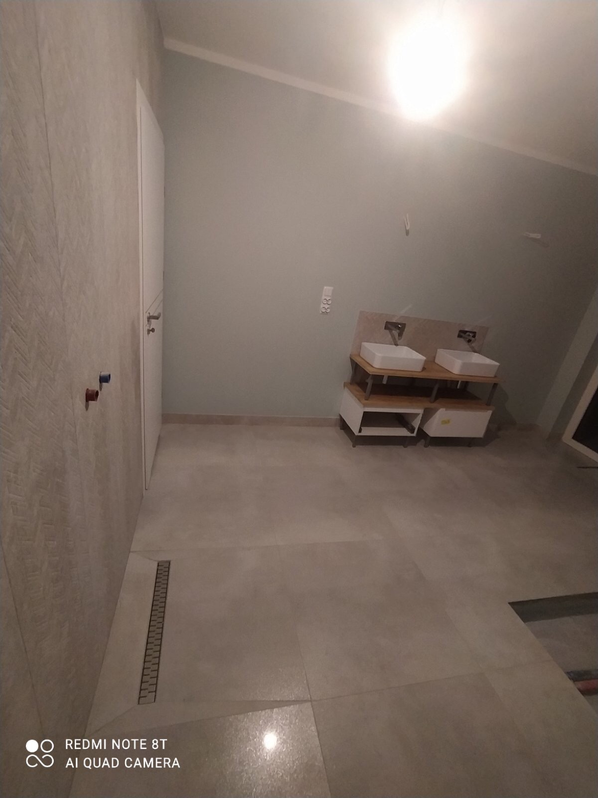 Nowoczesna łazienka w neutralnych barwach z podwójną umywalką na drewnianym stelażu i odpływem liniowym w podłodze, widoczne przyłącza wodne na ścianie.