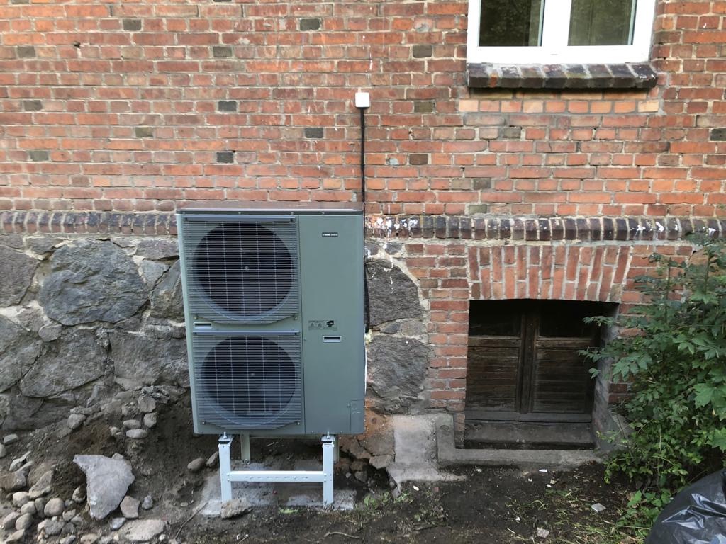 Szara pompa ciepła typu split zamontowana na białych wspornikach przed ścianą z czerwonej cegły i kamienia, z widocznym oknem i małym wejściem do piwnicy.