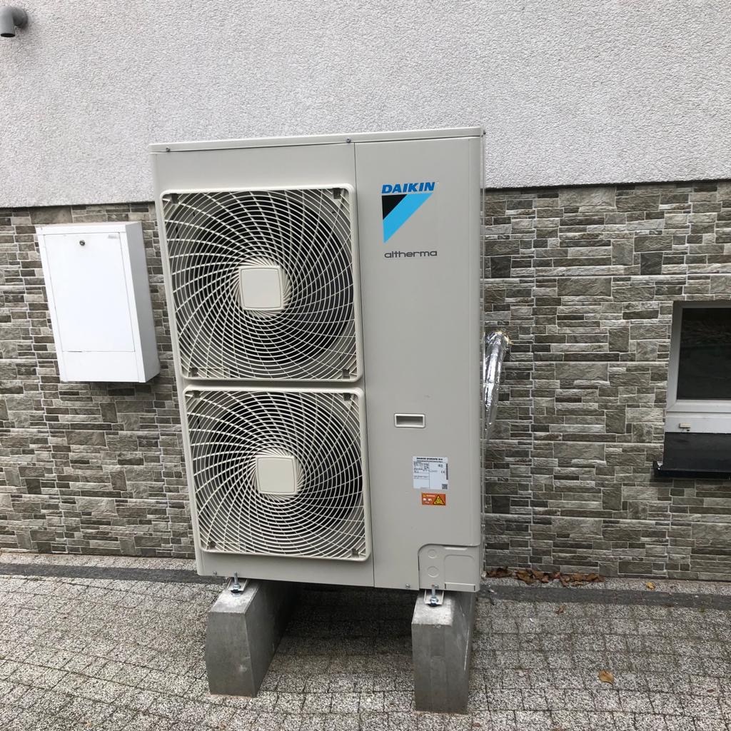 Zewnętrzna jednostka pompy ciepła Daikin Altherma zamontowana na betonowych bloczkach przy ścianie budynku z szarej cegły. Widoczne dwa wentylatory, biała skrzynka na ścianie i fragment okna.