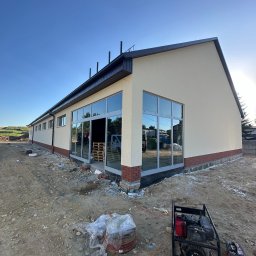 MAT-MAC MATEUSZ Krajewski - Budynek po tynkowaniu z dużymi oknami, widoczny generator prądu i teren budowy. Jasna elewacja, ceglana podmurówka i ciemny dach. Słoneczny dzień.