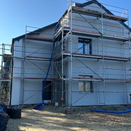 MAT-MAC MATEUSZ Krajewski - Dom w trakcie prac elewacyjnych, otoczony rusztowaniem. Widoczne worki na odpady i niebieskie rury wentylacyjne. Świeżo nałożony tynk w jasnym odcieniu. Błękitne niebo w tle.