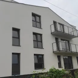 Nowoczesny budynek wielorodzinny z jasną, gładką elewacją, czarnymi oknami i balkonami z szklanymi balustradami, widok z dołu na tle pochmurnego nieba i słupa energetycznego.