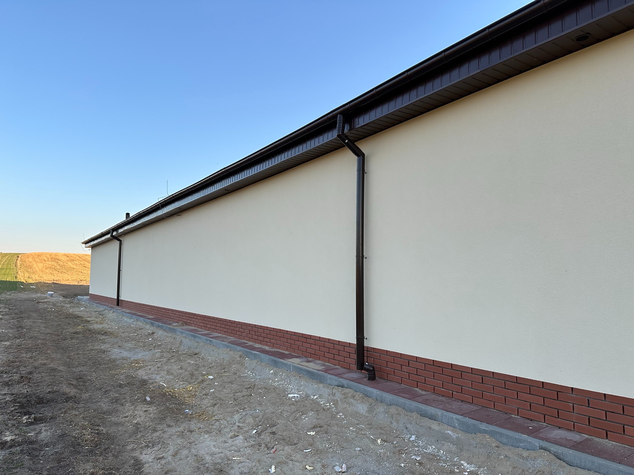 Elewacja budynku z jasnym tynkiem, brązową podmurówką z cegły klinkierowej i brązowymi rynnami. Widok z boku, w tle pole i błękitne niebo.