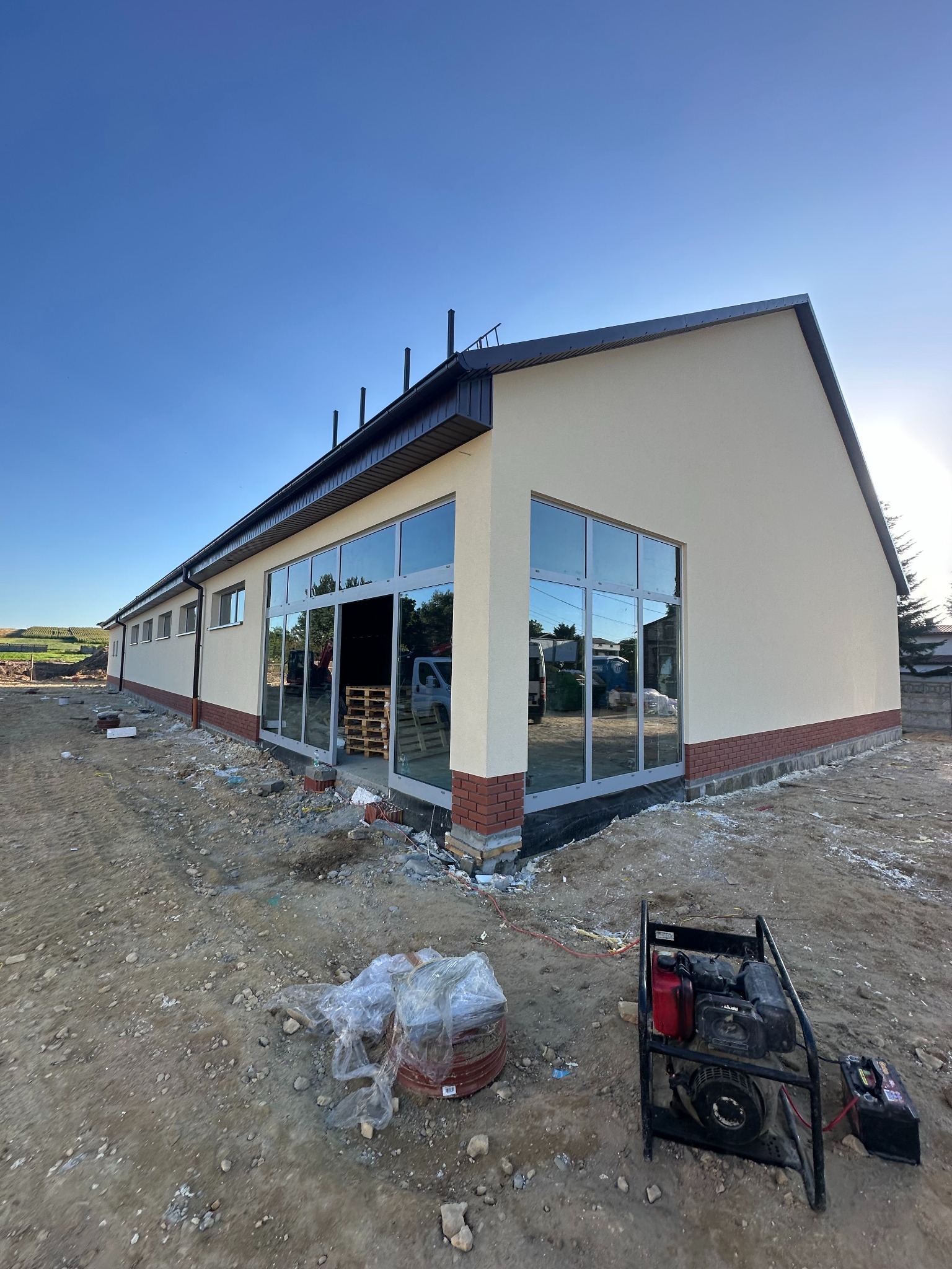 Budynek po tynkowaniu z dużymi oknami, widoczny generator prądu i teren budowy. Jasna elewacja, ceglana podmurówka i ciemny dach. Słoneczny dzień.