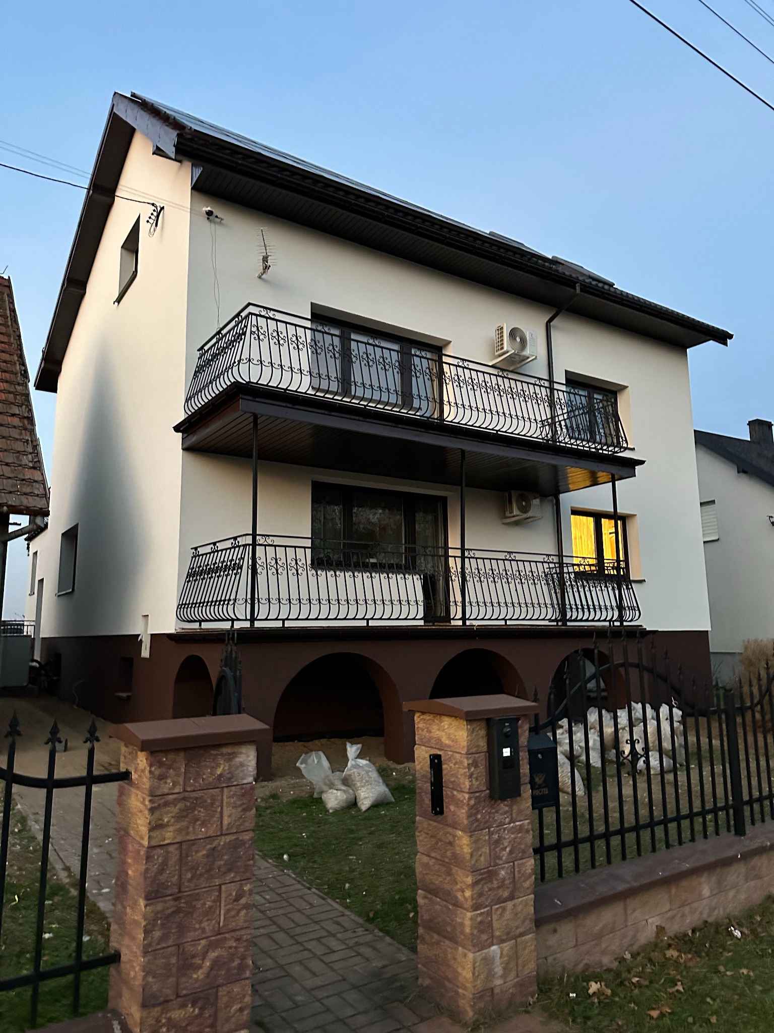 Dwupiętrowy dom z balkonami i klimatyzacją, po remoncie elewacji. Widoczne elementy dekoracyjne i ogrodzenie z ceglanymi słupkami. Ujęcie z dołu, w tle błękitne niebo.