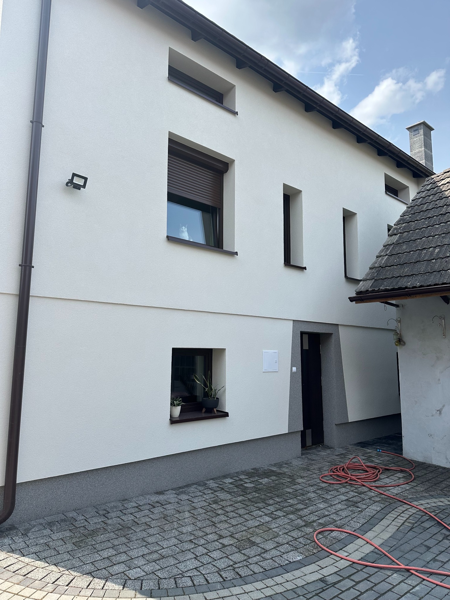 Biała elewacja dwukondygnacyjnego domu z brązowymi oknami i rynnami, widok z zewnątrz na kostkę brukową i częściowo zwinięty wąż ogrodowy.