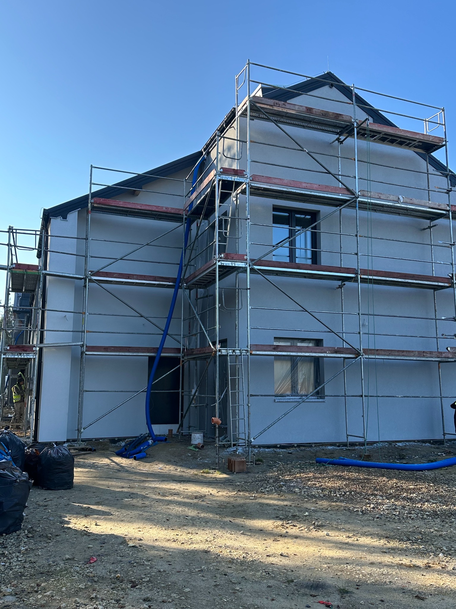 Dom w trakcie prac elewacyjnych, otoczony rusztowaniem. Widoczne worki na odpady i niebieskie rury wentylacyjne. Świeżo nałożony tynk w jasnym odcieniu. Błękitne niebo w tle.