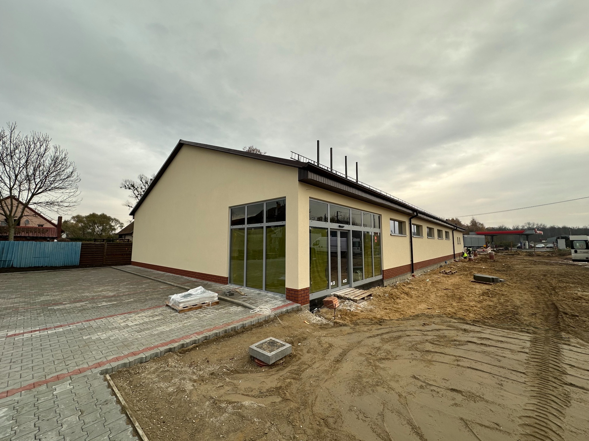 Budynek po renowacji elewacji z dużymi oknami i nową kostką brukową. Widoczne materiały budowlane i teren po pracach ziemnych. Pochmurne niebo w tle.